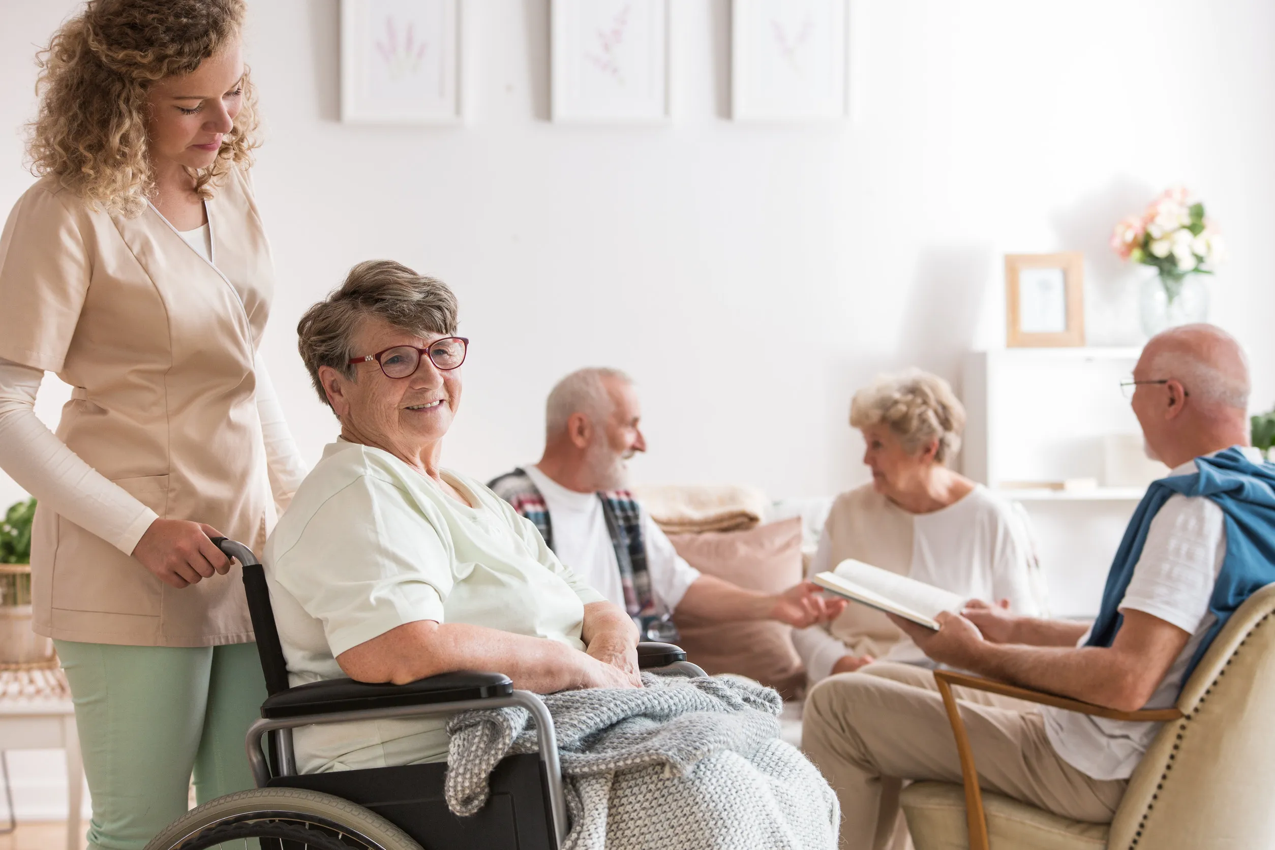 115417441_m Young beautiful caregiver and positive senior woman sitting at wheelchair in nursing home for elderly